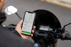 Delivery driver holding a smartphone with a navigation and order notification screen next to a scooter during a food delivery shift.