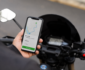 Delivery driver holding a smartphone with a navigation and order notification screen next to a scooter during a food delivery shift.